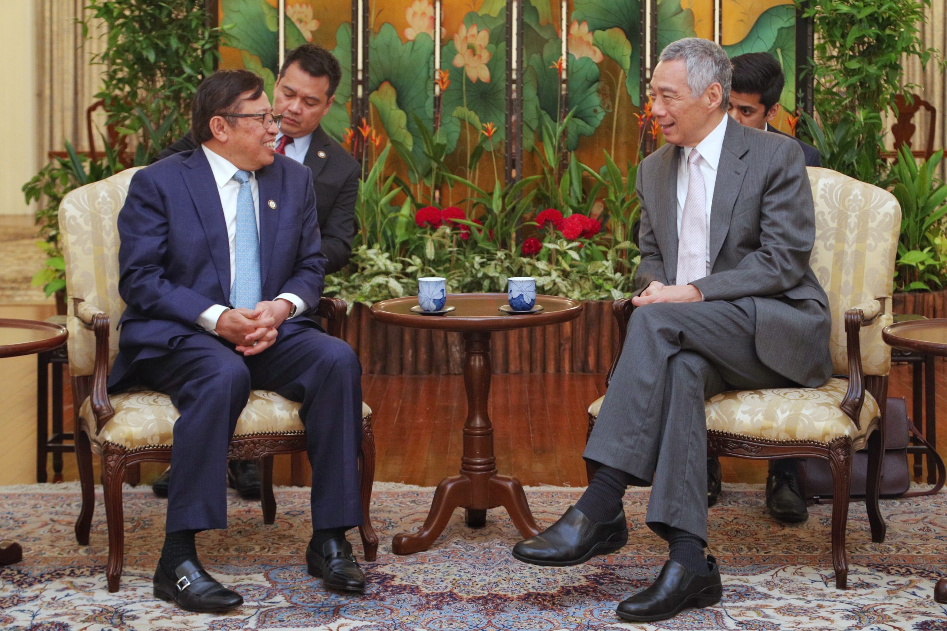 Two men in suits sit facing each other in ornate chairs with a small table between them.